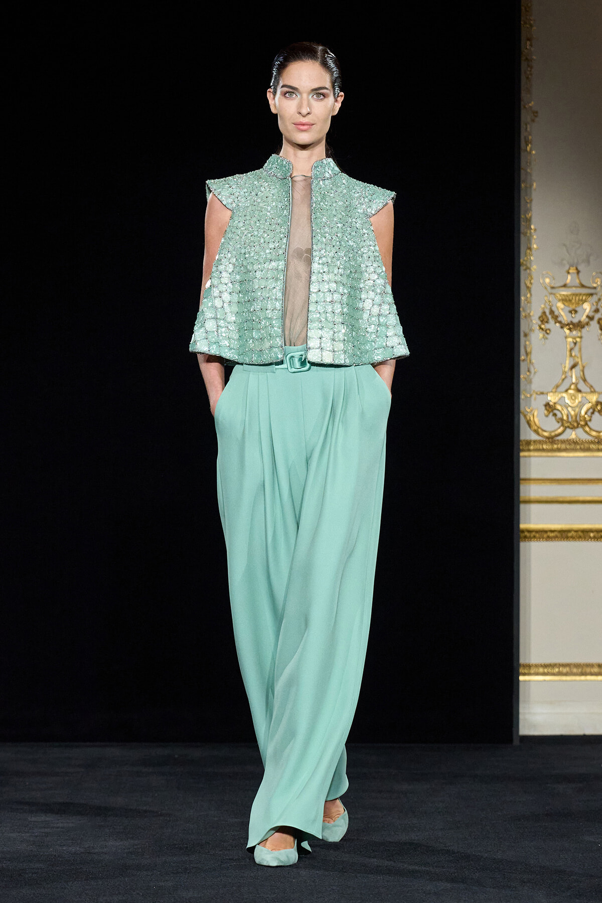 Model walks a runway in a mint green, beaded sleeveless vest over a sheer taupe blouse and high-waist wide-leg pants with a matching belt.
