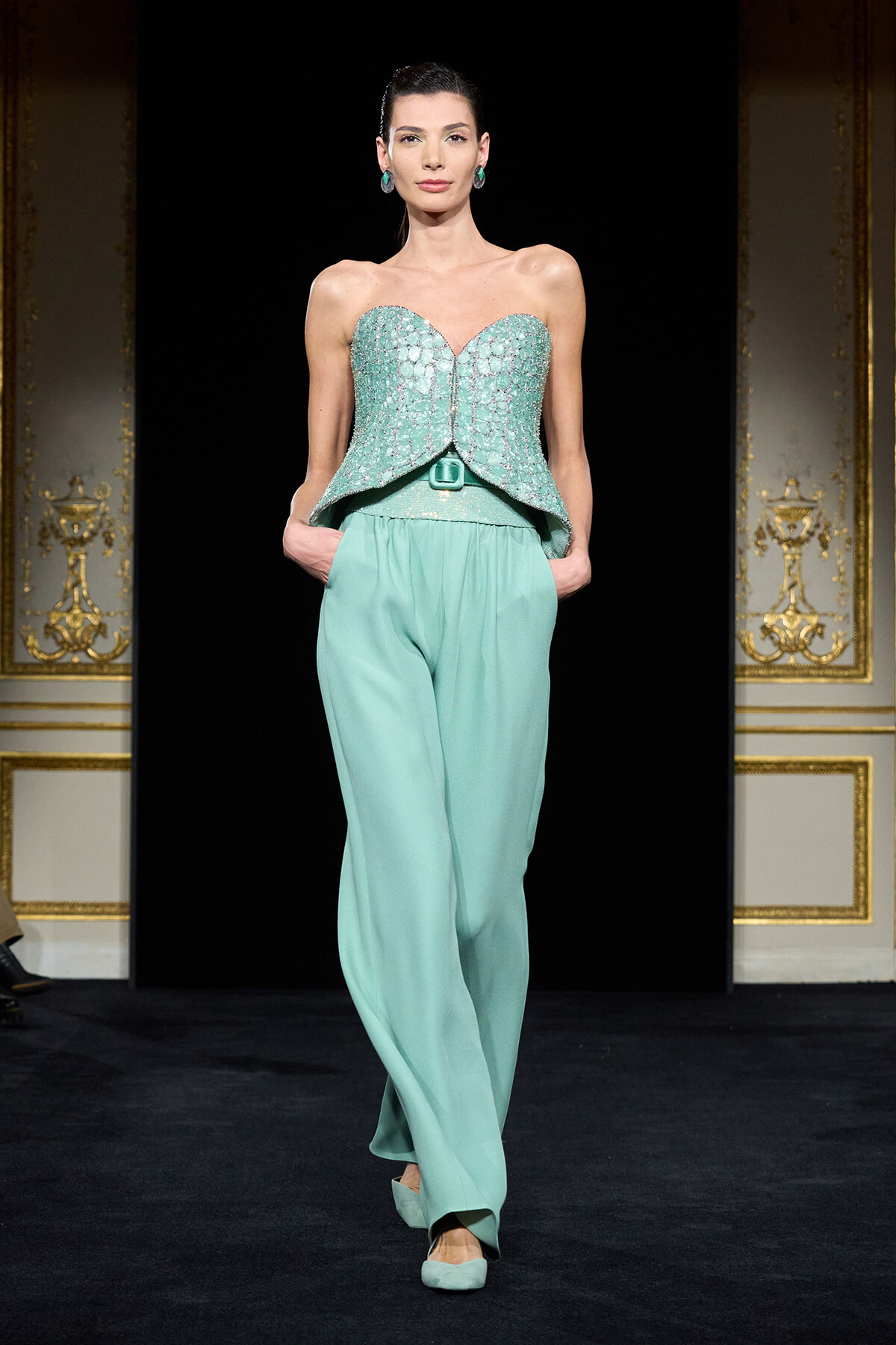 Model on a runway wearing a turquoise strapless beaded peplum top and wide-leg pants with matching flats.
