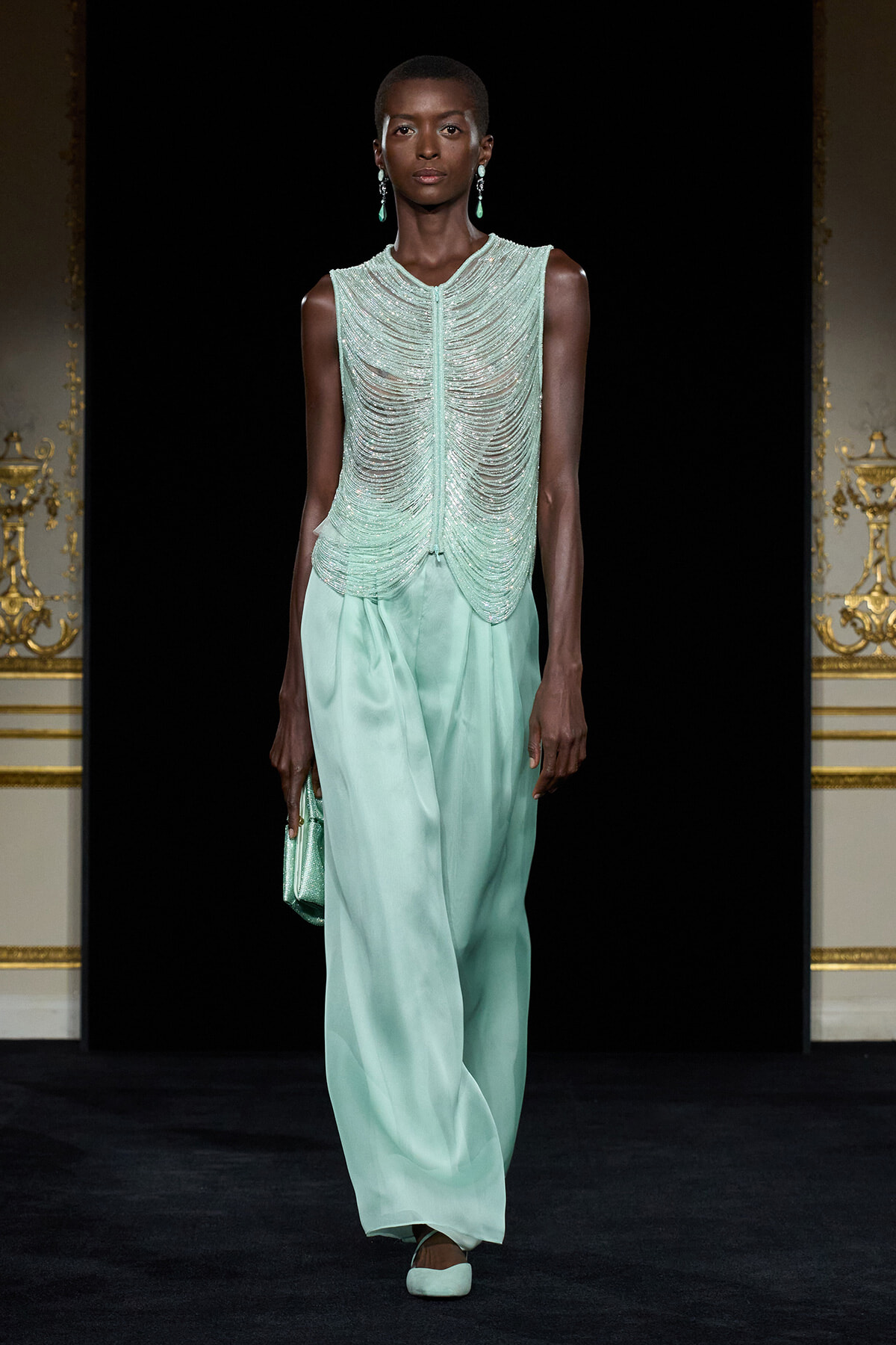 Model on a runway wearing a mint-green beaded sleeveless top with cascading beadwork and matching wide-leg satin pants, carrying a mint clutch.