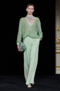 Model on a runway wearing a mint-green sequined V-neck sweater over a sheer pale gray collared blouse, with wide mint pants and a matching clutch in hand.
