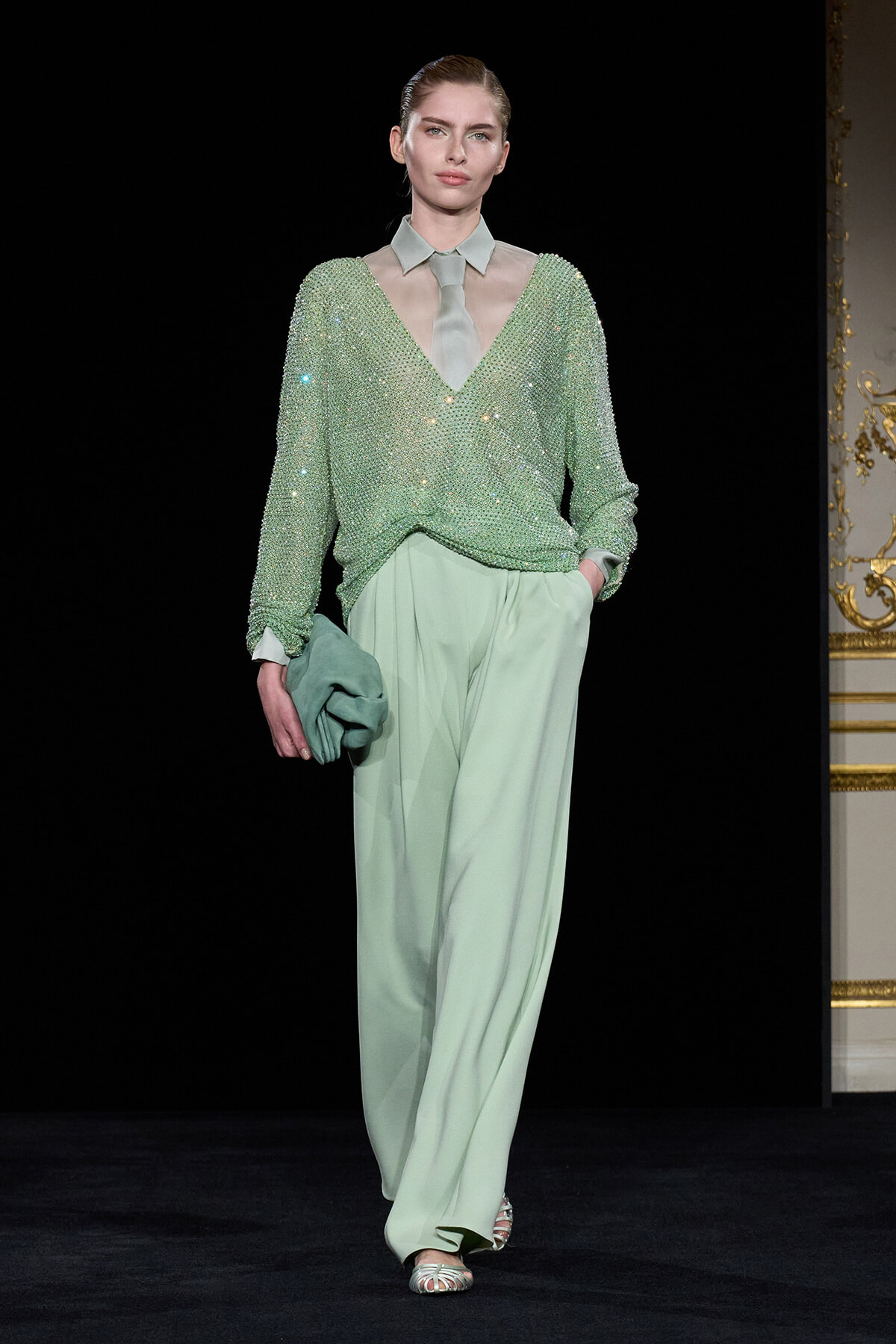 Model on a runway wearing a mint-green sequined V-neck sweater over a sheer pale gray collared blouse, with wide mint pants and a matching clutch in hand.