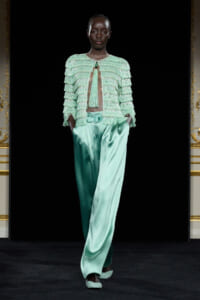 Runway model in mint-green fringe jacket with satin wide-leg pants and matching belt, walking on a black backdrop.