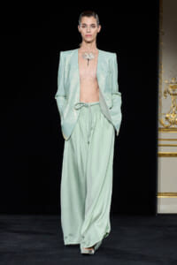 Model on a runway wearing a mint-green sequin blazer, sheer top, and matching wide-leg pants with a statement necklace.