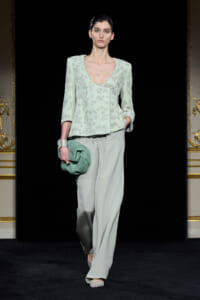Model walks a fashion runway wearing a pale mint lace top with wide V-neck and pale gray wide-leg trousers, clutching a green bag.