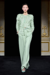Model walks the runway in a mint-green, tiered, ruffled top paired with wide-leg pants; hands in pockets on a black backdrop.