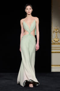 Model walking a runway in a pale mint green gown with a deep V bodice, cutouts at the waist, and beaded detailing, complemented by a long flowing skirt and fringe on the side.