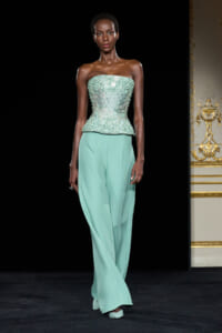 Model walking the runway in a strapless mint-green beaded corset top and matching wide-leg pants against a black backdrop.