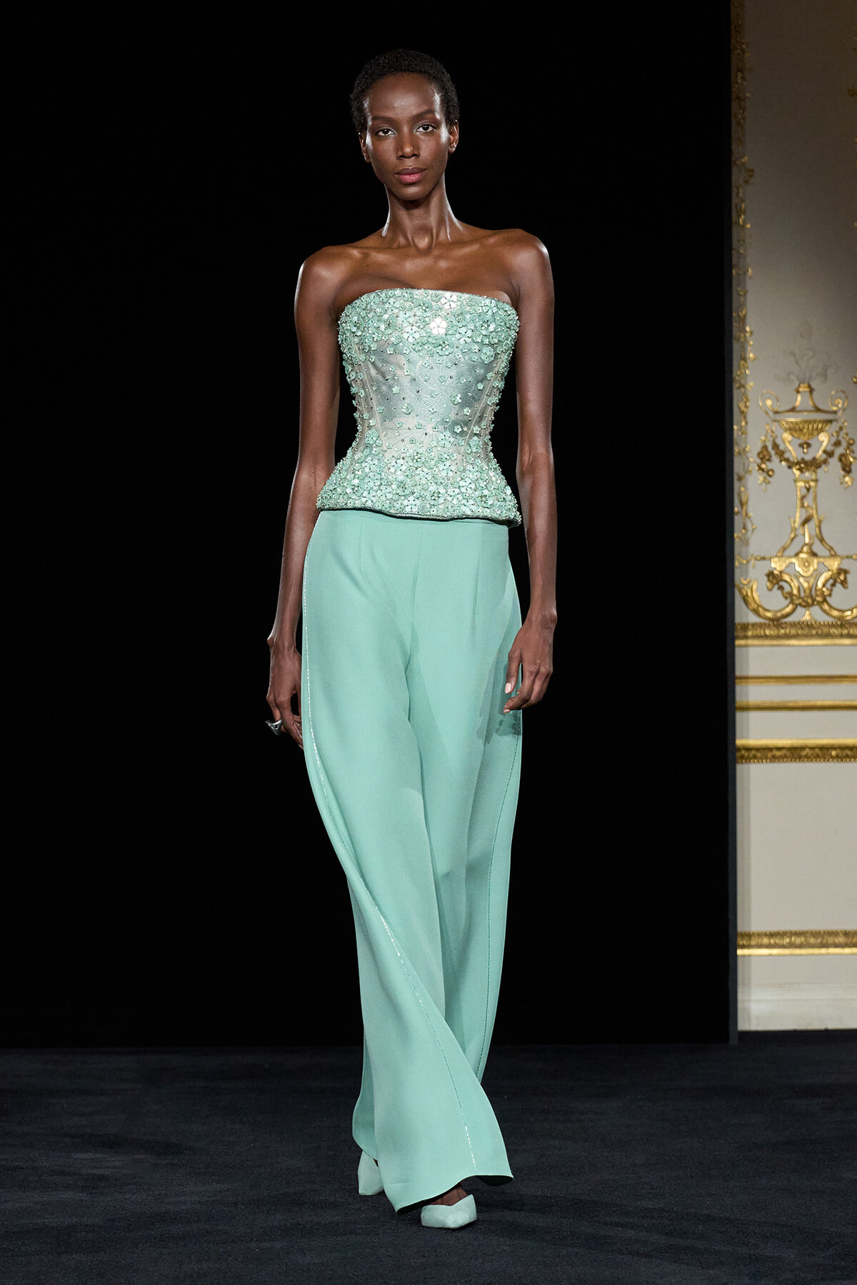 Model walking the runway in a strapless mint-green beaded corset top and matching wide-leg pants against a black backdrop.