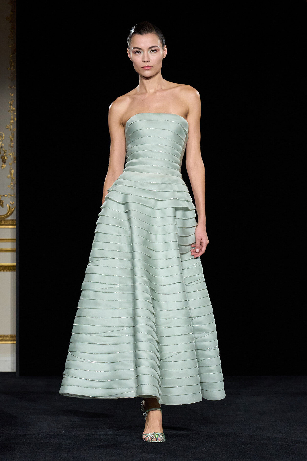 Model on a runway wearing a strapless, pale mint-green ballgown with horizontal banded layers and a voluminous tiered skirt, paired with silver sandals.