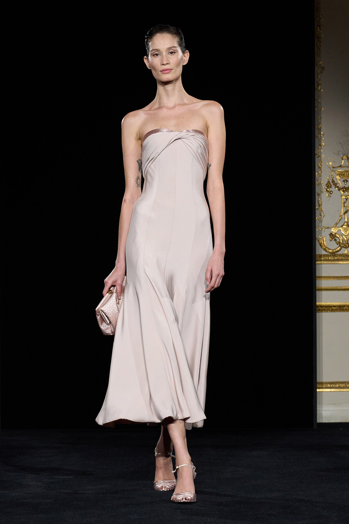 Model walks a runway in a pale pink strapless gown, holding a matching clutch and wearing delicate heels.
