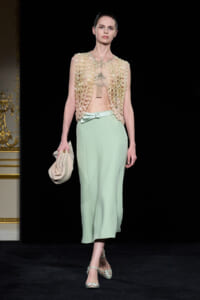 Model on a runway wearing a pale mint-green midi skirt with a beige beaded sleeveless top, holding a beige clutch and wearing strappy mint heels.