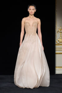 Model walking the runway in a strapless champagne gown with a beaded sweetheart bodice and flowing layered skirt.