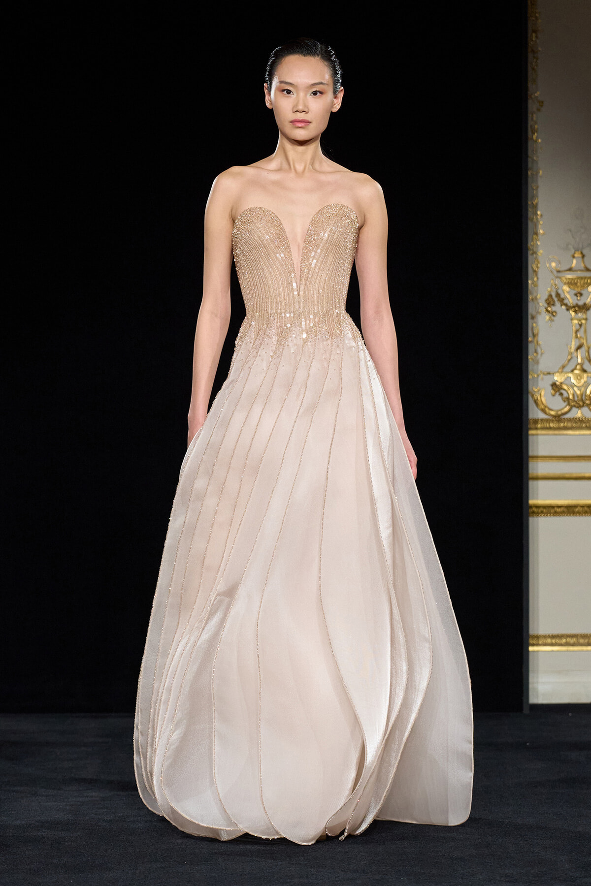 Model walking the runway in a strapless champagne gown with a beaded sweetheart bodice and flowing layered skirt.