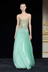Model on a runway wearing a mint green evening gown with a jeweled, sheer-embellished bodice and flowing pleated skirt