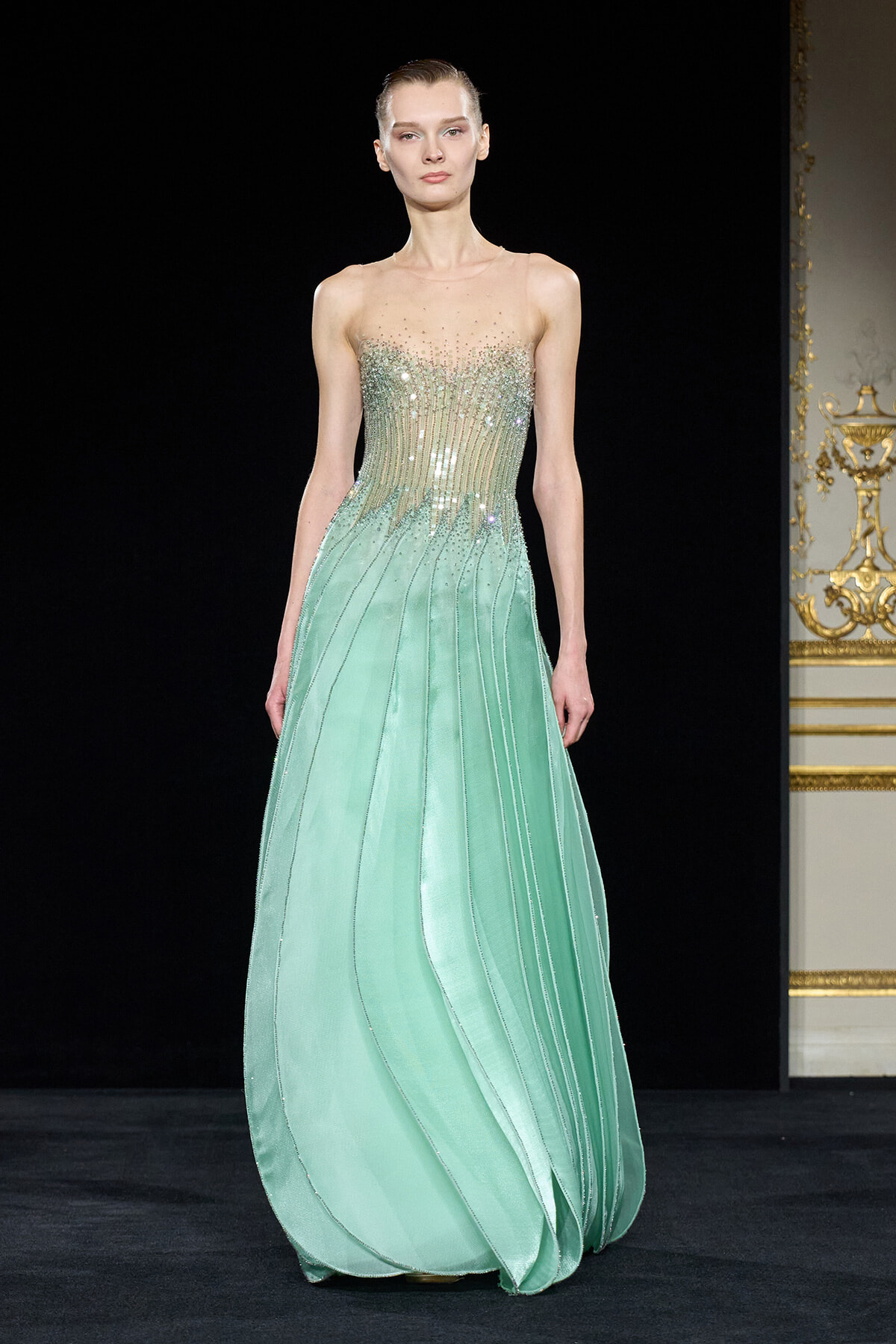 Model on a runway wearing a mint green evening gown with a jeweled, sheer-embellished bodice and flowing pleated skirt