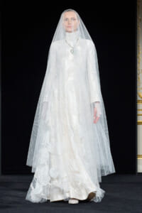 Model wearing a full-length white wedding gown with a long sheer veil on a runway stage with a dark backdrop.
