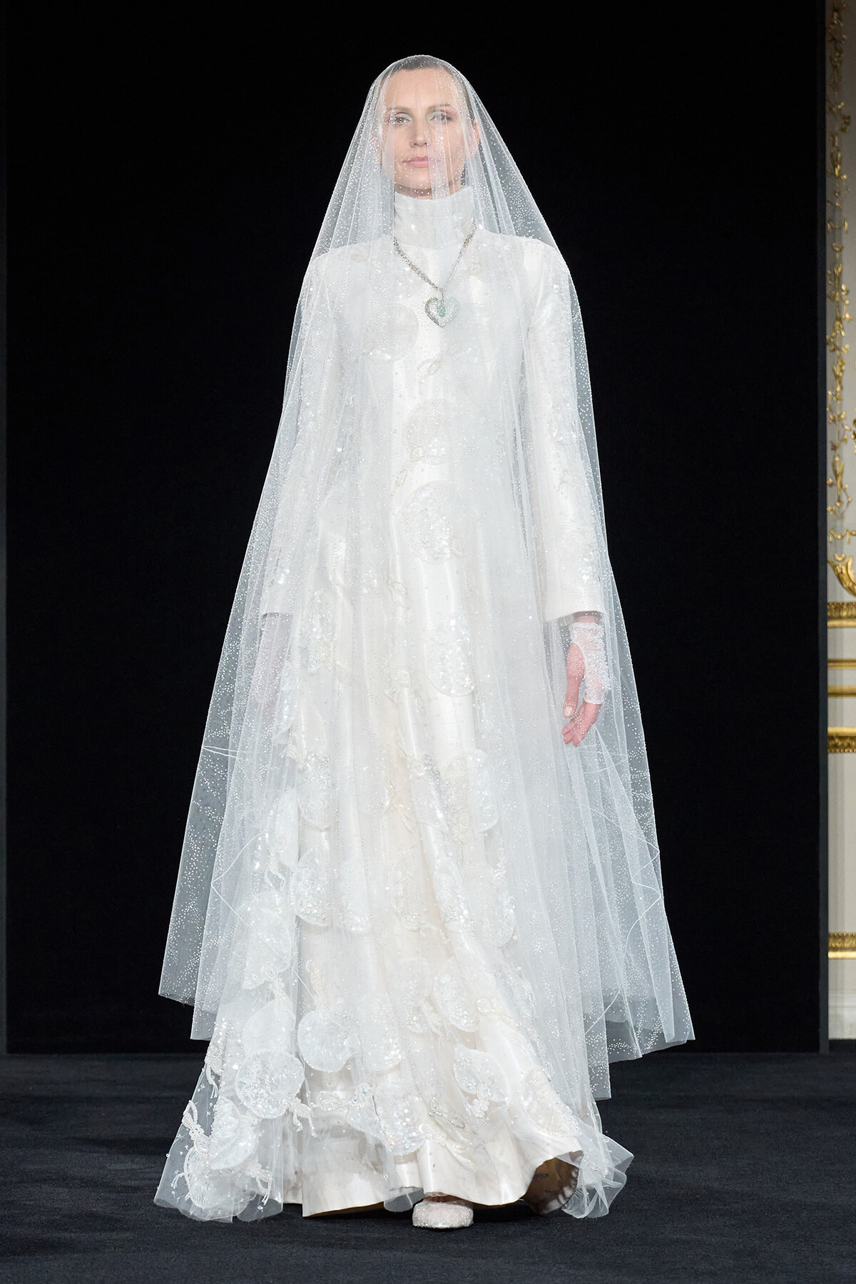 Model wearing a full-length white wedding gown with a long sheer veil on a runway stage with a dark backdrop.