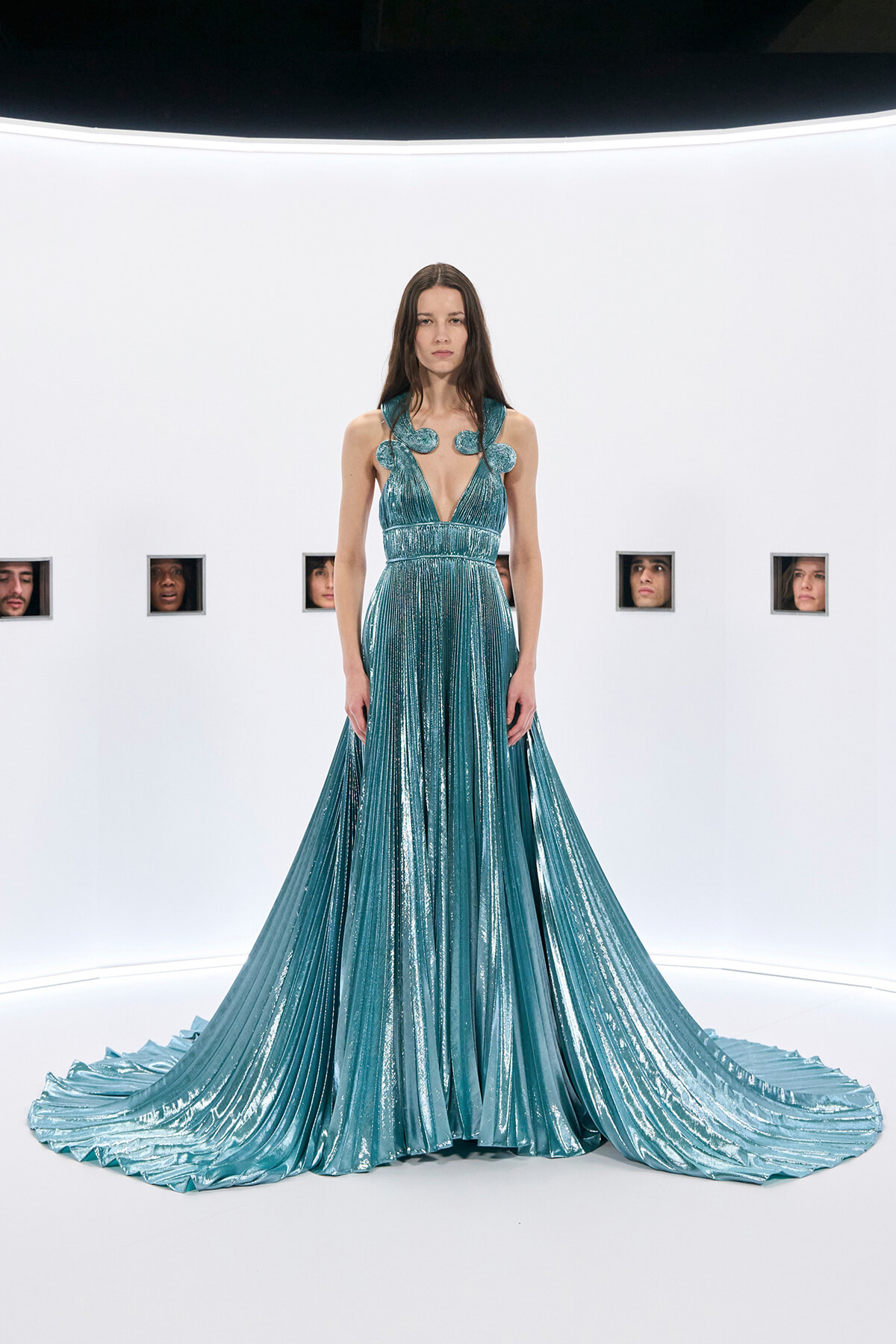 Model in a shimmering teal gown with a deep V-neck, decorative shoulder flaps, and a long pleated train on a white runway backdrop.