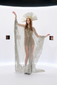 Model wearing a silver beaded gown with a lace cape and a gold sunburst halo in a white studio space.