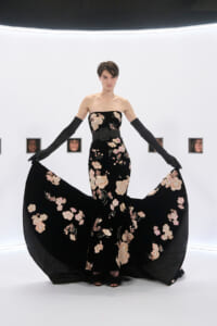 Model in a strapless black gown with pink floral embroidery, a wide belt, and long black gloves, lifting the skirt to reveal a flowing cape-like train.