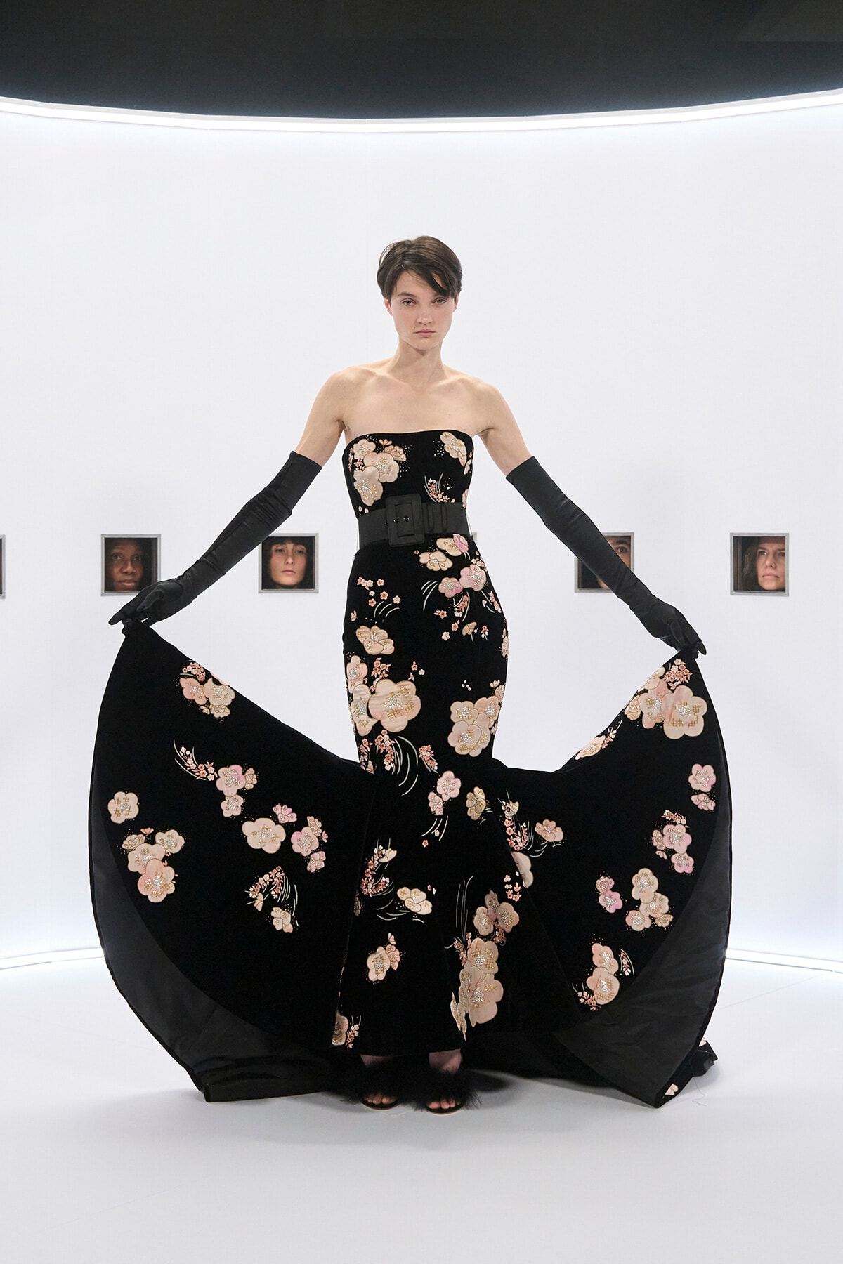 Model in a strapless black gown with pink floral embroidery, a wide belt, and long black gloves, lifting the skirt to reveal a flowing cape-like train.