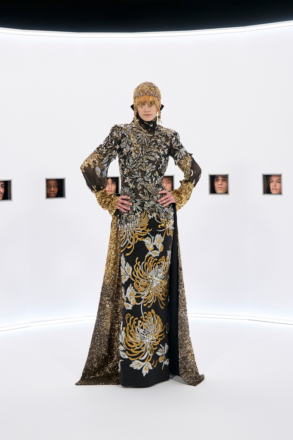 Model in an elaborate black gown with gold and silver embroidery, standing with hands on hips in a white studio; head is adorned with a gold headdress and fringe. Full-length fashion photo.