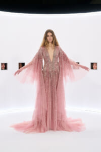 Model in a sparkling pink gown with long sheer cape sleeves, arms out, in a bright white studio set.