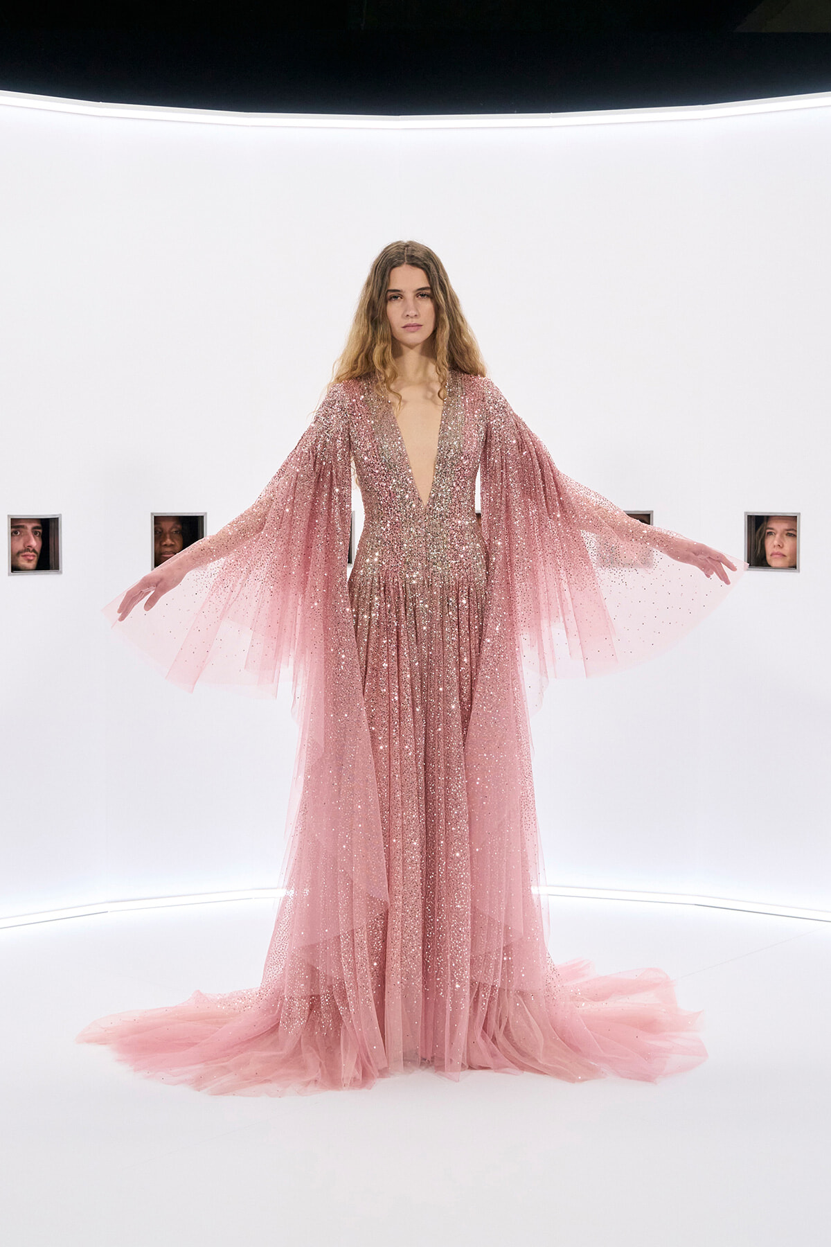 Model in a sparkling pink gown with long sheer cape sleeves, arms out, in a bright white studio set.