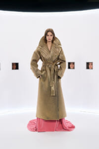 Model in a metallic gold oversized trench coat with wide lapels, belted at the waist, standing on pink fabric at the hem in a white studio.