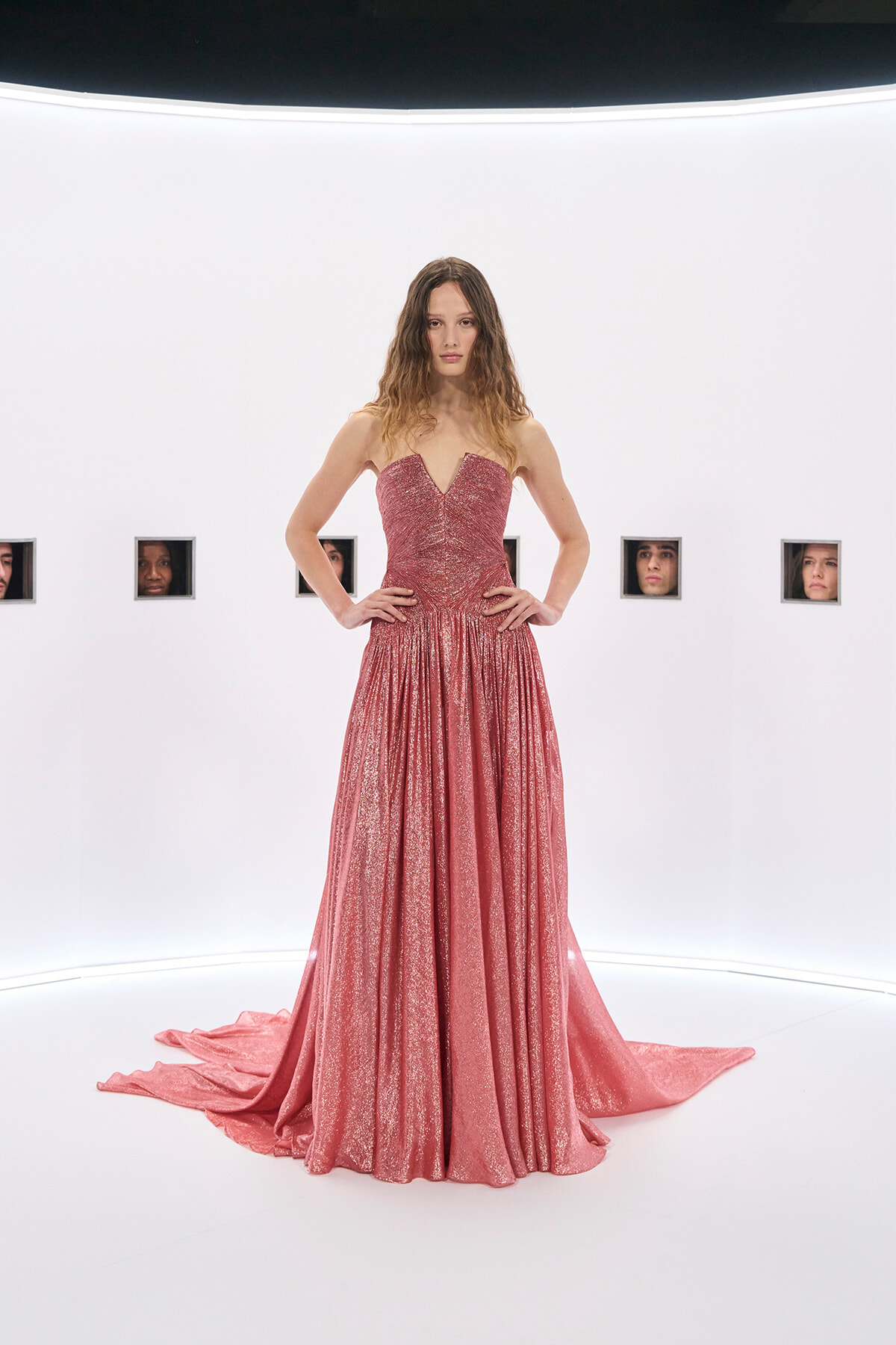 Model wearing a sparkling pink strapless gown with a long train, standing against a white studio backdrop.