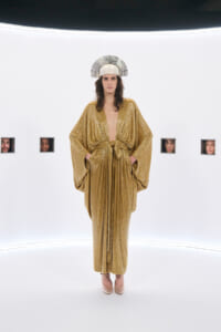 Model wearing a gold, sequined kimono-style gown with a tied waist, standing in a white gallery space with small framed faces on the wall and a sunburst headpiece on their head.