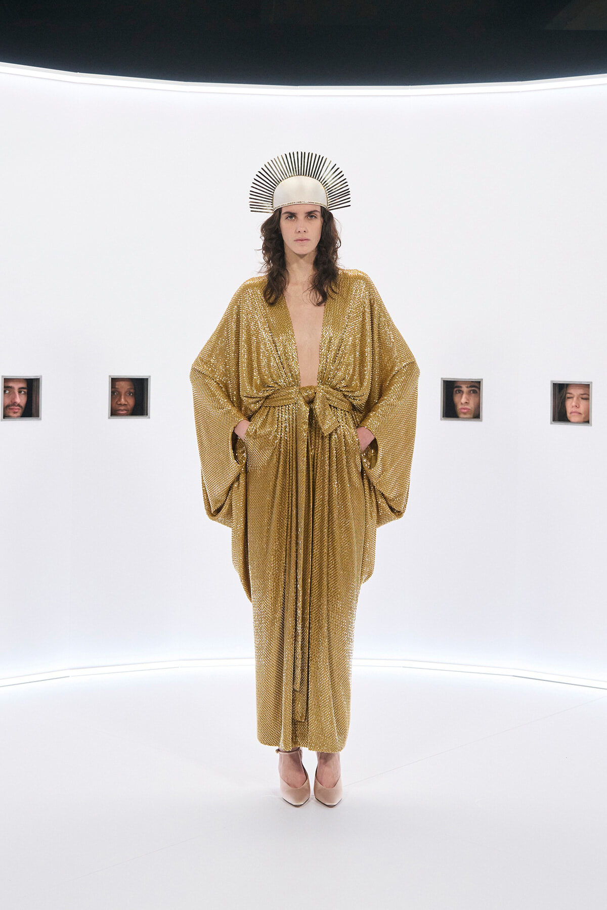 Model wearing a gold, sequined kimono-style gown with a tied waist, standing in a white gallery space with small framed faces on the wall and a sunburst headpiece on their head.