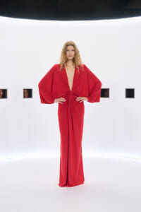 Model in a red, floor-length gown with voluminous cape sleeves stands with hands on hips in a bright white gallery.