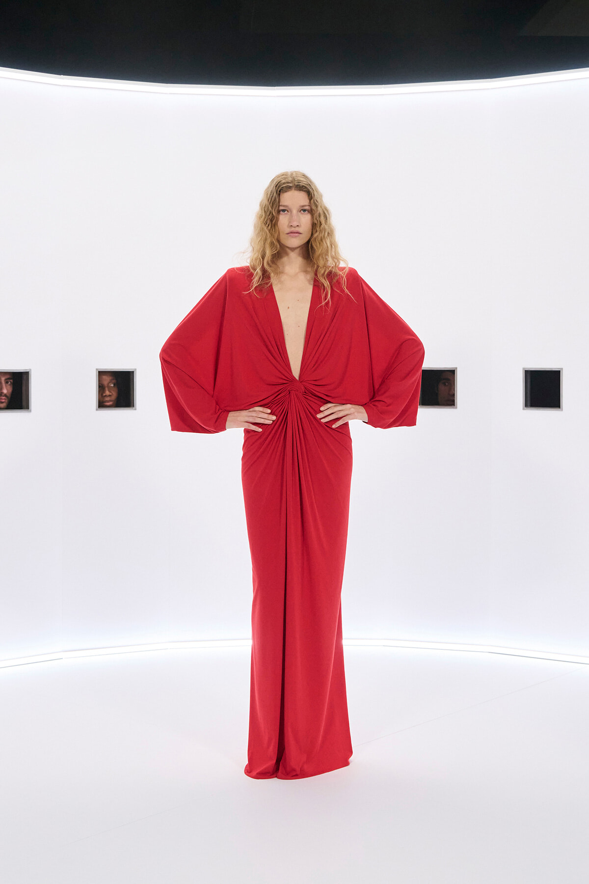 Model in a red, floor-length gown with voluminous cape sleeves stands with hands on hips in a bright white gallery.