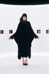 Model wearing a long black velvet cape dress with a sparkling collar, standing in a stark white studio with small windowed faces in the background.