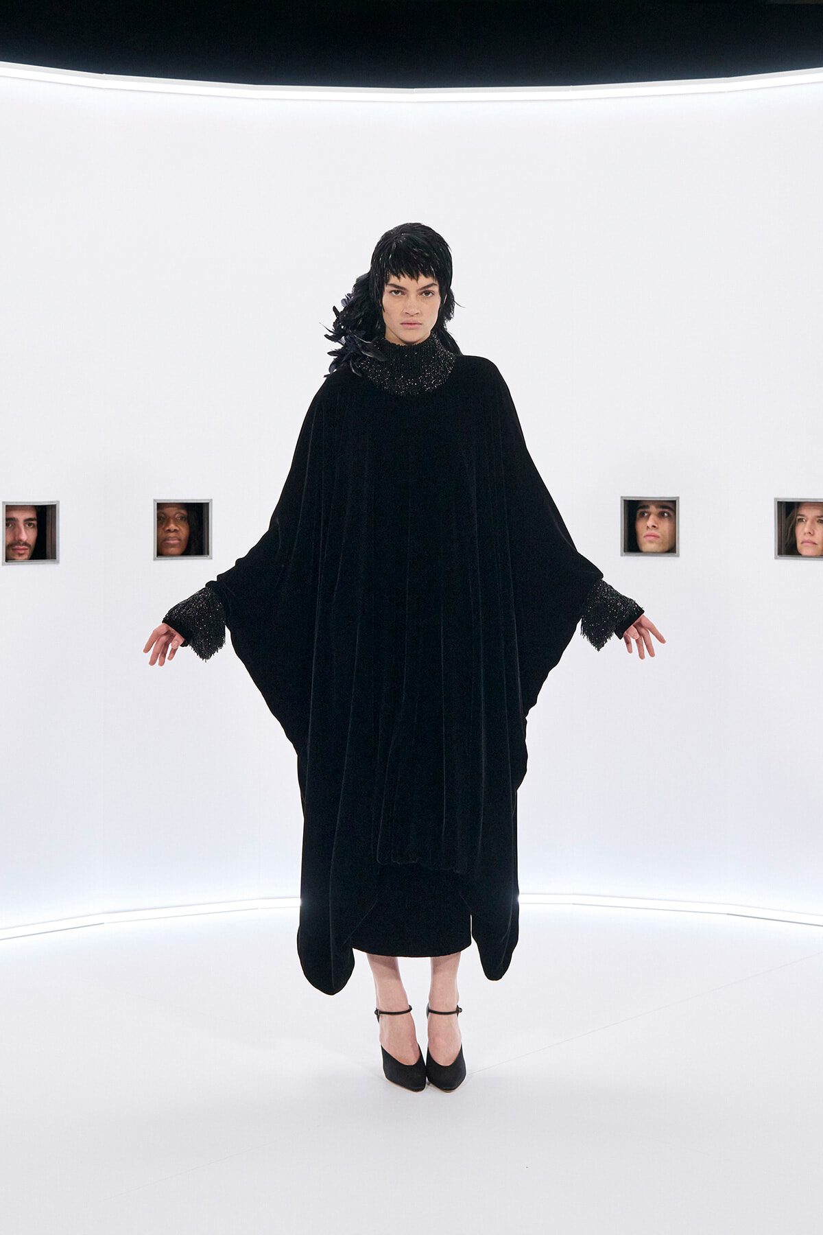 Model wearing a long black velvet cape dress with a sparkling collar, standing in a stark white studio with small windowed faces in the background.