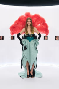Model wearing a teal and black avant-garde gown with a gold bodice and a large red feather cape in a white studio setting with small framed faces in the background
