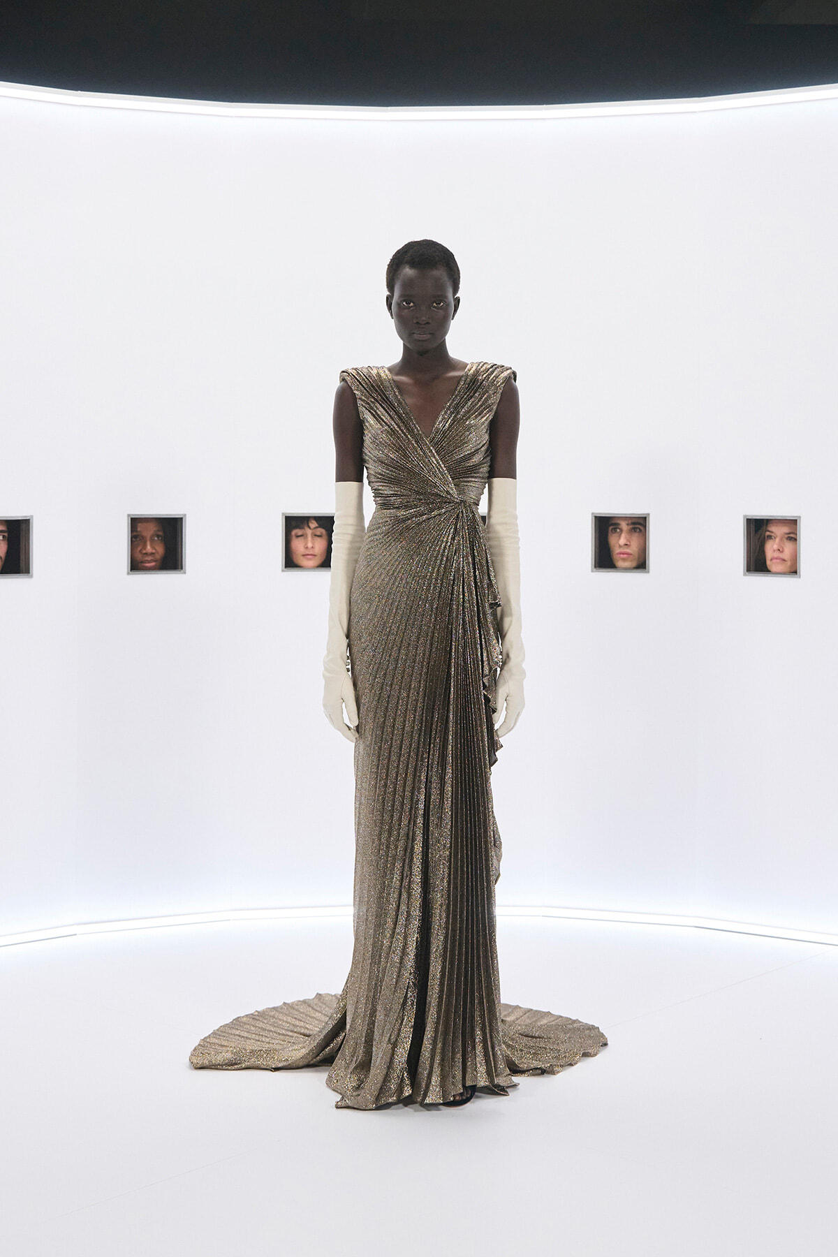 Black female model wearing a shimmering gold wrap gown with long cream gloves, standing in a stark white studio with small window panels showing faces behind her.