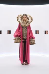 Model in a magenta cape with a large gold ruff collar and matching gold cuffs, standing in a white studio.