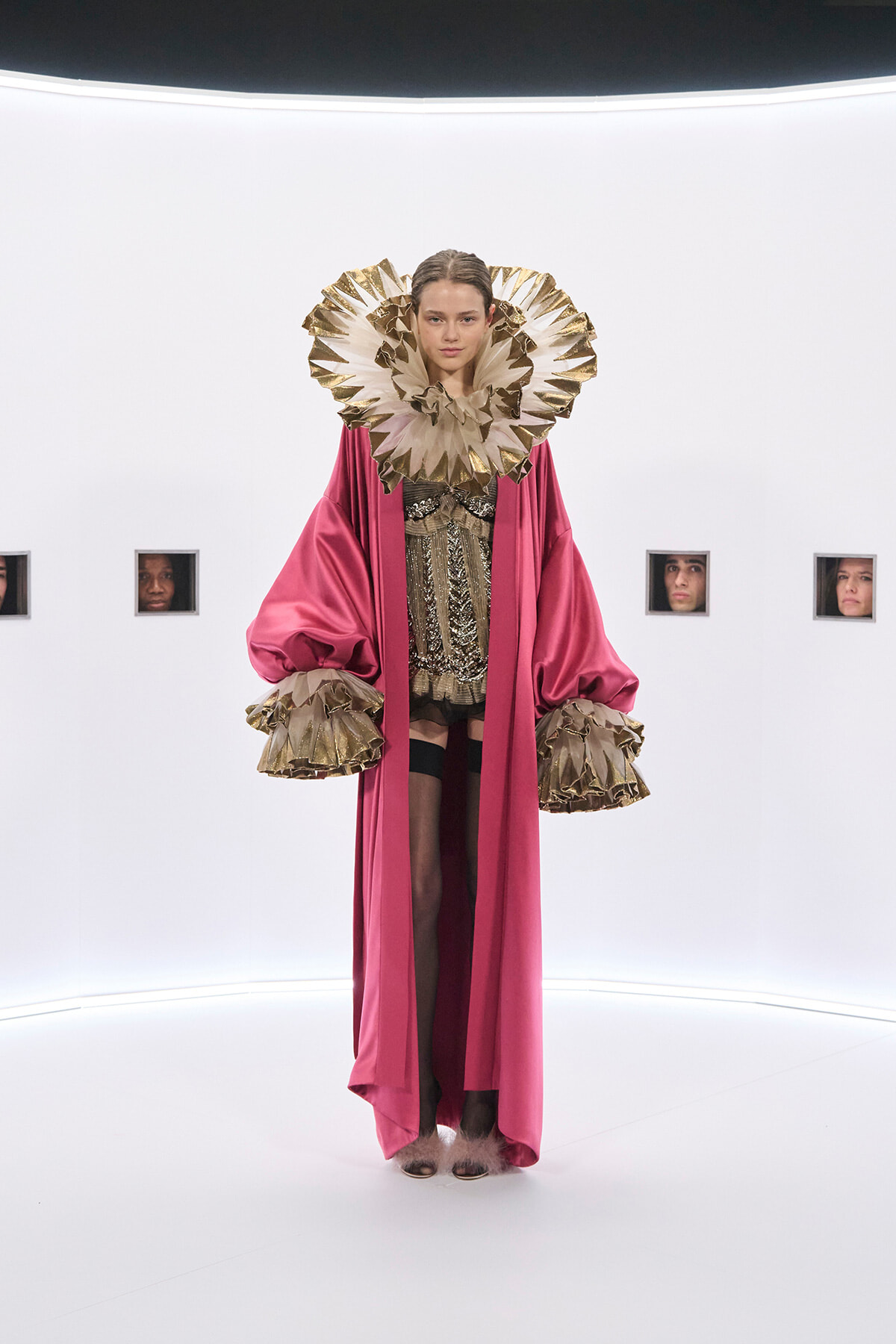 Model in a magenta cape with a large gold ruff collar and matching gold cuffs, standing in a white studio.
