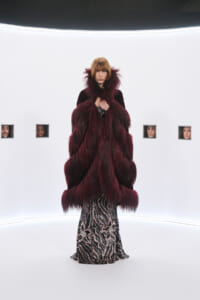 Full-length shot of a model wearing a voluminous burgundy fur coat over a black-and-silver zebra-patterned gown, posed in a stark white studio runway setting.