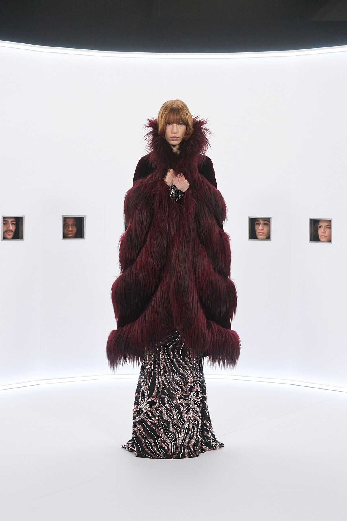 Full-length shot of a model wearing a voluminous burgundy fur coat over a black-and-silver zebra-patterned gown, posed in a stark white studio runway setting.