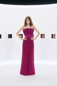 Model wearing a magenta strapless gown with a pleated bodice and wide belt, standing with hands on hips in a white studio setting.