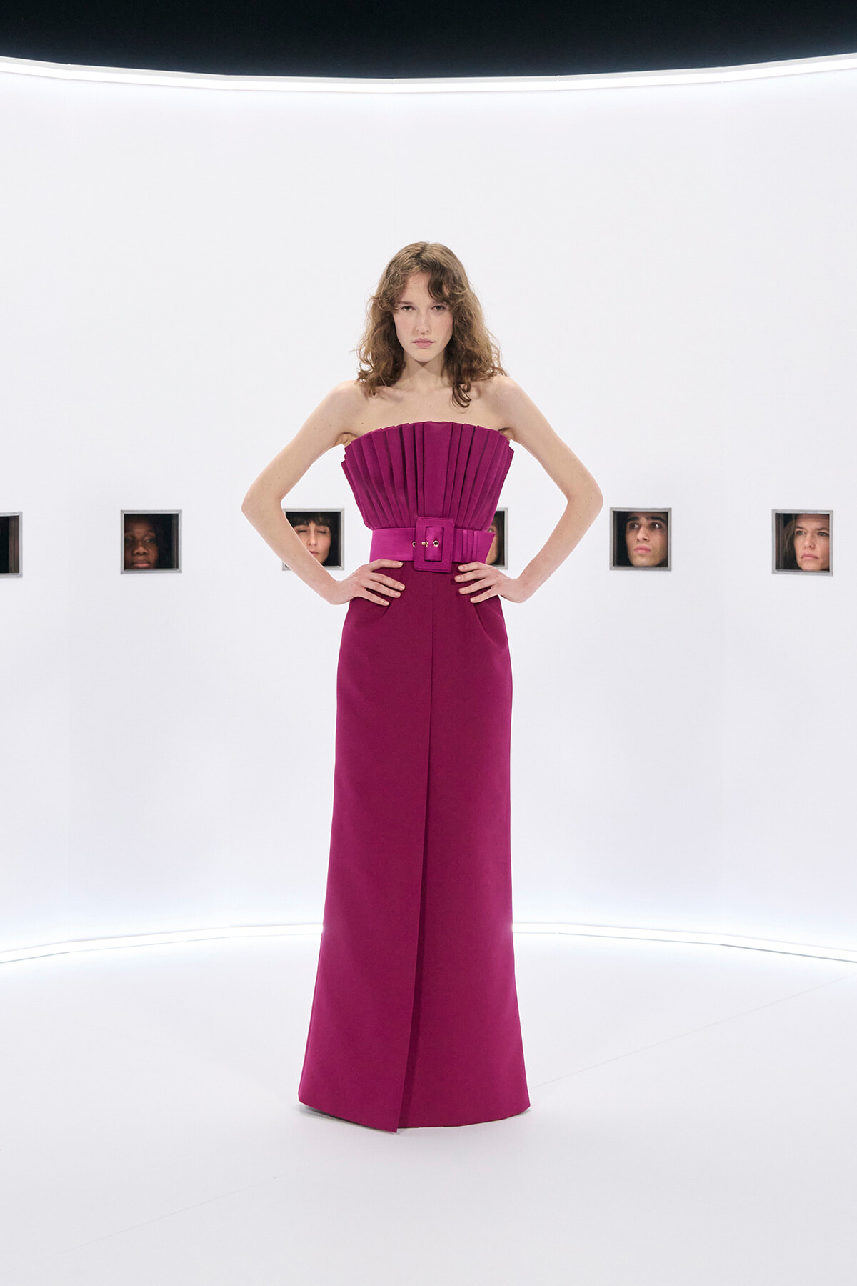 Model wearing a magenta strapless gown with a pleated bodice and wide belt, standing with hands on hips in a white studio setting.