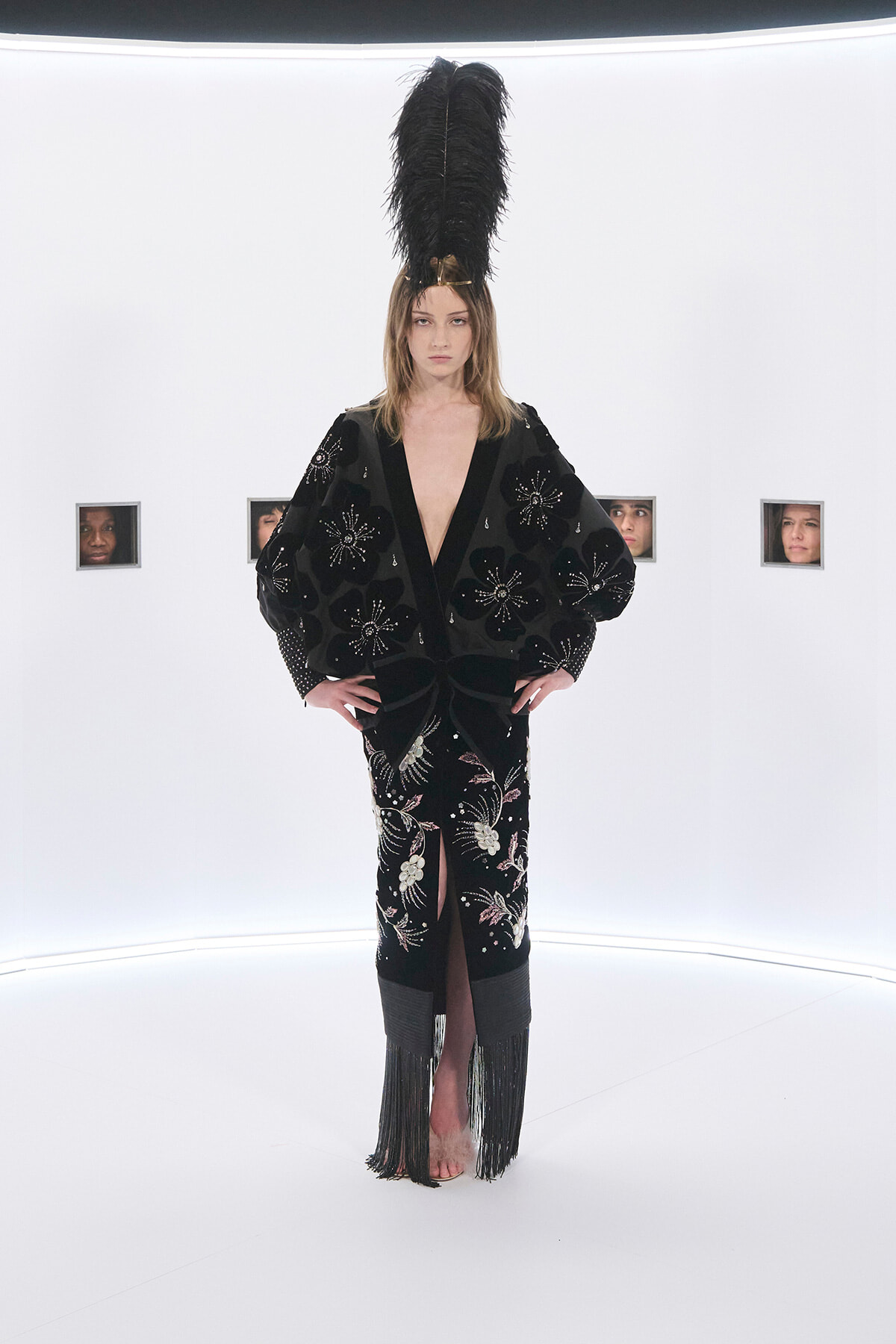 Model on a white studio runway wearing a black embellished robe with floral embroidery and a tall black feather headpiece, hands on hips, barefoot.