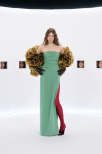 Model poses in a strapless green gown with dramatic gold feathered sleeves, black gloves, red tights, and black heeled shoes in a white studio backdrop with small portrait windows on the wall.