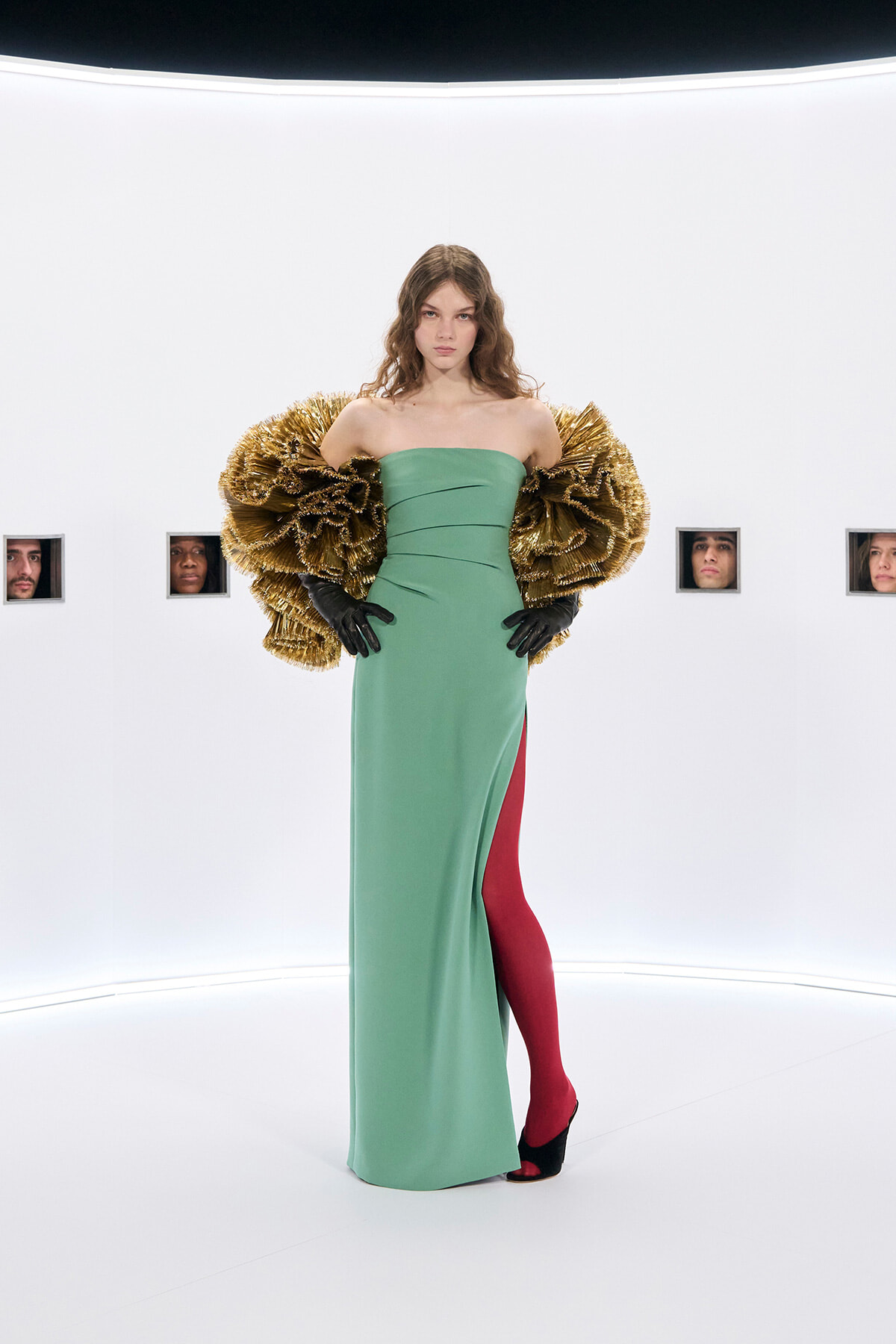 Model poses in a strapless green gown with dramatic gold feathered sleeves, black gloves, red tights, and black heeled shoes in a white studio backdrop with small portrait windows on the wall.