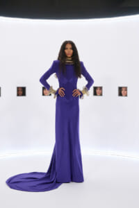 Model in a regal royal‑purple gown with gold neck and wrist jewelry stands with hands on hips in a white gallery, small framed portraits behind her.