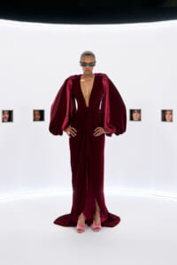 Model wearing a deep red velvet gown with oversized sleeves, posing in a stark white studio with small framed faces on the wall.