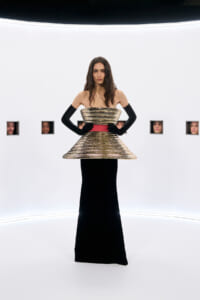 Model in a gold sculptural strapless top and long black gown with a red waistband, wearing black gloves, standing in a white studio with small square windows showing faces